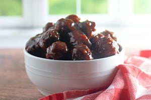 A white bowl filled with Slow Cooker Hot Honey Meatballs sits on a wooden surface next to a red and white checkered cloth, with a blurred window in the background.