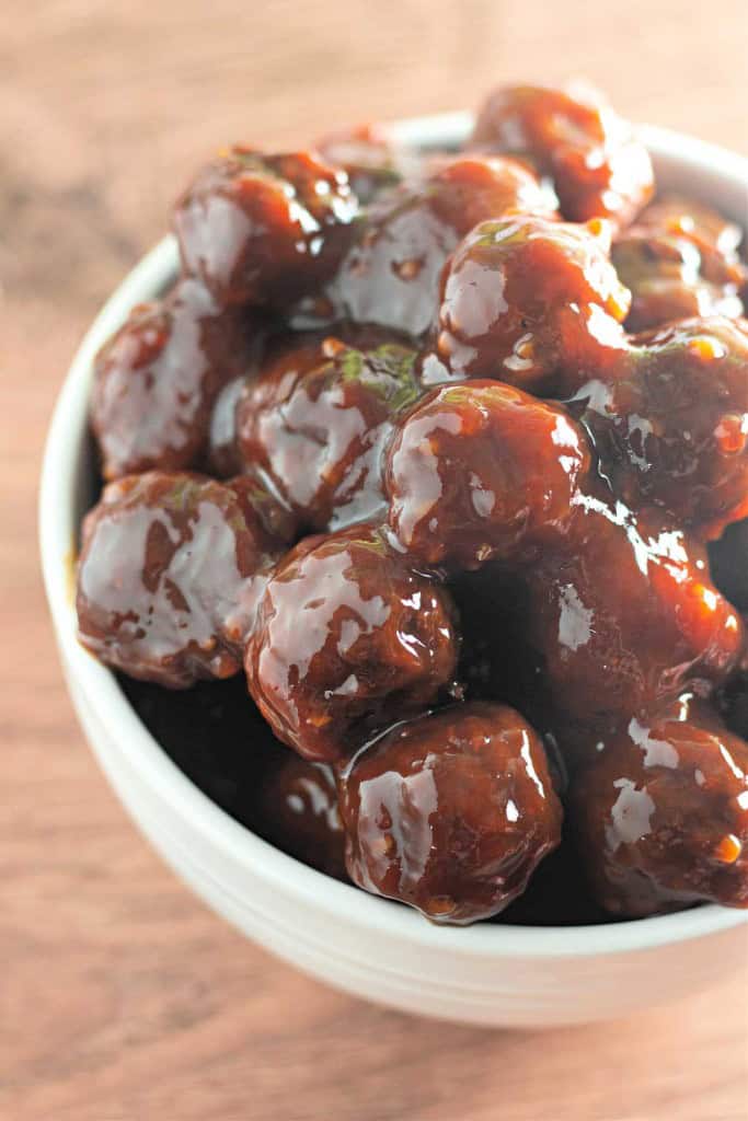A white bowl filled with Slow Cooker Hot Honey Meatballs, glazed and coated in a thick, dark brown sauce, sits on a wooden surface.
