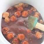 Slow Cooker Hot Honey Meatballs are covered in sauce inside a slow cooker, with a green spatula partially submerged.