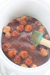 Slow Cooker Hot Honey Meatballs are covered in sauce inside a slow cooker, with a green spatula partially submerged.