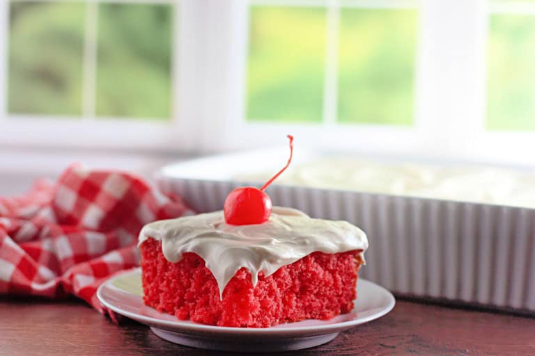 A slice of maraschino cherry cake with white frosting and a cherry on top sits on a plate, with a dish of cake and a red checked cloth in the background.