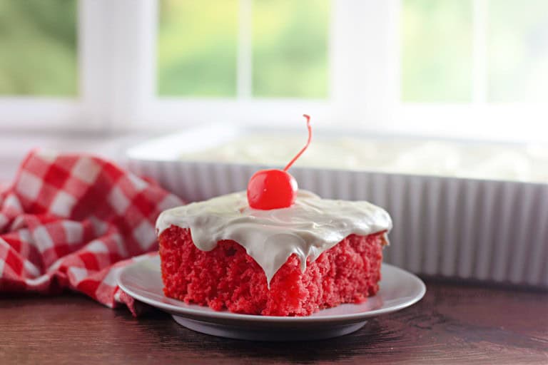 A slice of maraschino cherry cake with white frosting and a cherry on top sits on a white plate, with a baking dish and a red checkered cloth in the background.