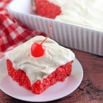A square slice of bright red maraschino cherry cake with white frosting and a cherry on top sits on a white plate, next to a baking dish and a red checkered cloth.