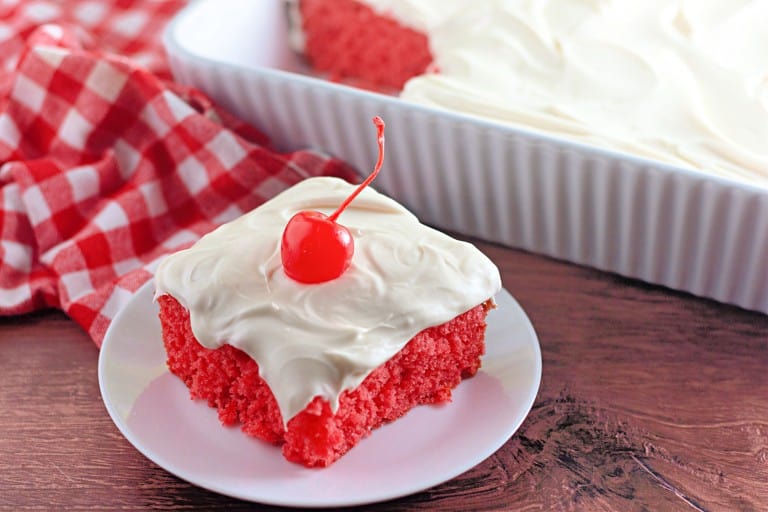 A square slice of bright red maraschino cherry cake with white frosting and a cherry on top sits on a white plate, next to a baking dish and a red checkered cloth.