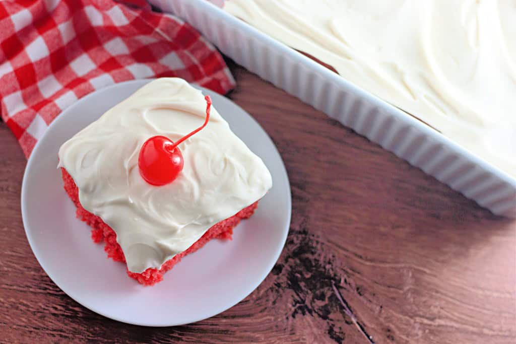 A slice of maraschino cherry cake with white frosting and a cherry on top sits on a plate, beside a baking dish filled with more of the same cake.