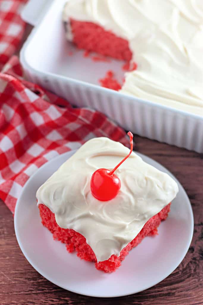 A square piece of pink cake with white frosting and a cherry on top sits on a plate; the rest of the cake is in a baking dish in the background.