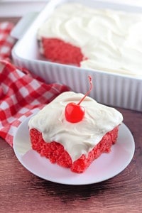 A slice of bright red cake with white frosting and a cherry on top sits on a white plate, with the remaining cake in a baking dish in the background.