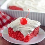 A slice of bright red maraschino cherry cake with white frosting and a cherry on top sits on a plate, with a larger dish of the same cake in the background.