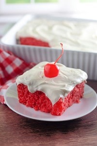 A slice of bright red maraschino cherry cake with white frosting and a cherry on top sits on a plate, with a larger dish of the same cake in the background.