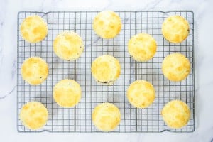 Fourteen golden muffins cool on a metal wire rack, arranged in a grid pattern on a white marble surface.
