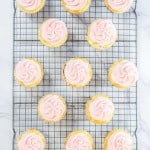 Fourteen cupcakes with pink frosting sit on a metal cooling rack placed on a white marble surface.