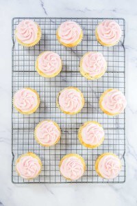 Fourteen cupcakes with pink frosting sit on a metal cooling rack placed on a white marble surface.