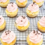 Twelve cupcakes with pink frosting and chocolate chips sit on a wire cooling rack over a marble surface.