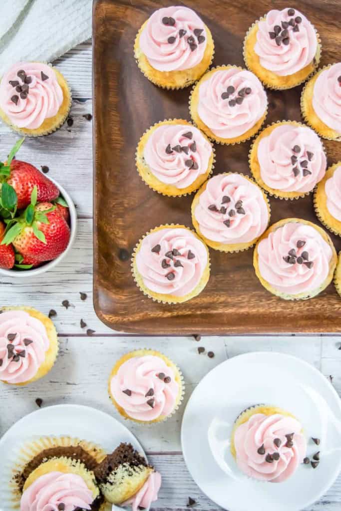 A wooden tray and plates with vanilla cupcakes topped with pink frosting and chocolate chips, next to a bowl of strawberries on a white wooden surface.