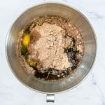 A mixing bowl containing eggs, cocoa powder, liquid, and other ingredients, ready to be mixed on a marble surface.