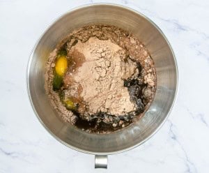 A mixing bowl containing eggs, cocoa powder, liquid, and other ingredients, ready to be mixed on a marble surface.
