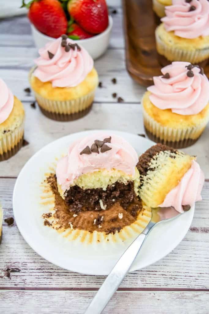 A cupcake with pink frosting, partially eaten to reveal chocolate and vanilla layers, sits on a white plate with a spoon. Other cupcakes and strawberries are in the background.