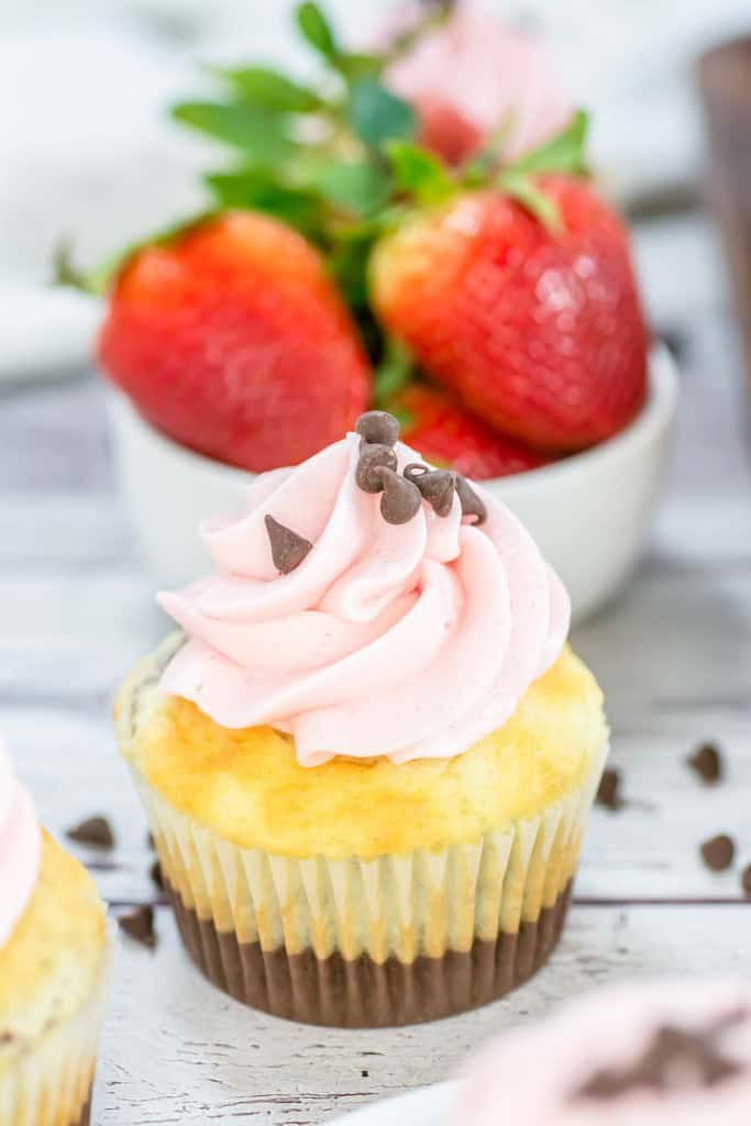 A vanilla cupcake with pink frosting and chocolate chips sits in front of a bowl of fresh strawberries on a white wooden surface.