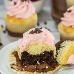 A cupcake with pink frosting, chocolate sprinkles, and a visible chocolate center sits on a plate with a fork. Other cupcakes and strawberries are in the background.