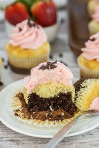 A cupcake with pink frosting, chocolate sprinkles, and a visible chocolate center sits on a plate with a fork. Other cupcakes and strawberries are in the background.