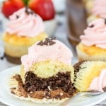 A cupcake with pink frosting is cut in half, revealing a chocolate center. Other similarly decorated cupcakes and strawberries are visible in the background.