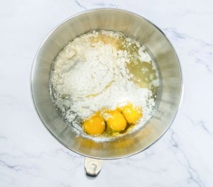 A metal mixing bowl containing flour, three egg yolks, and liquid ingredients on a marble countertop.