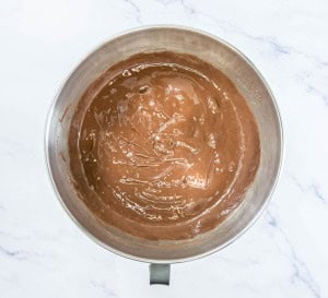 A stainless steel mixing bowl filled with smooth chocolate cake batter on a white marble surface.