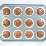A muffin tin lined with paper cups, each filled with chocolate batter, is placed on a white marble surface.