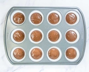 A muffin tin lined with paper cups, each filled with chocolate batter, is placed on a white marble surface.
