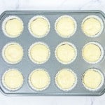 A muffin tin lined with paper cups, each filled with pale cupcake batter, rests on a white marble surface.