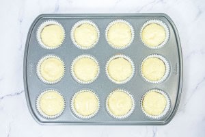 A muffin tin lined with paper cups, each filled with pale cupcake batter, rests on a white marble surface.