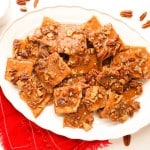 Rectangular pieces of pecan praline toffee are arranged on a white oval plate, with pecans scattered around and a red cloth napkin underneath.