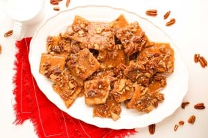 Rectangular pieces of pecan praline toffee are arranged on a white oval plate, with pecans scattered around and a red cloth napkin underneath.