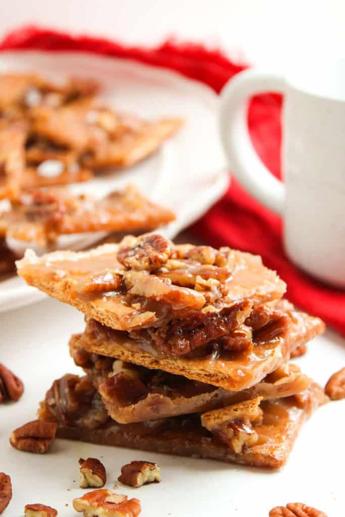 A stack of pecan toffee brittle pieces sits on a white surface, with scattered pecan pieces, a plate of more brittle, and a white mug in the background.