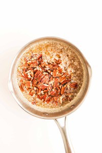 A stainless steel saucepan containing melted butter and chopped pecans on a white background.