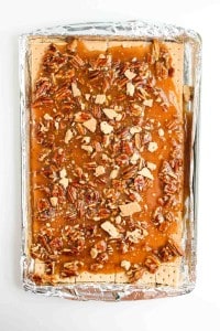 A baking sheet lined with foil holds a layer of graham crackers topped with chopped pecans and caramel sauce.