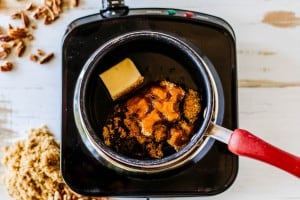 A saucepan with butter, brown sugar, and syrup for pecan pie dip sits on an electric stove, with spilled brown sugar and chopped nuts nearby on a white surface.