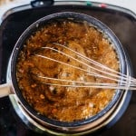 A pot of bubbling brown pecan pie dip is being stirred with a metal whisk on a stovetop.