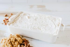 A white rectangular baking dish filled with a creamy white pecan pie dip sits on a light surface, with brown sugar and pecans nearby.