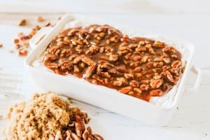 A white baking dish filled with a creamy pecan pie dip, topped with a caramelized pecan mixture, sits next to a pile of brown sugar and pecans on a white surface.