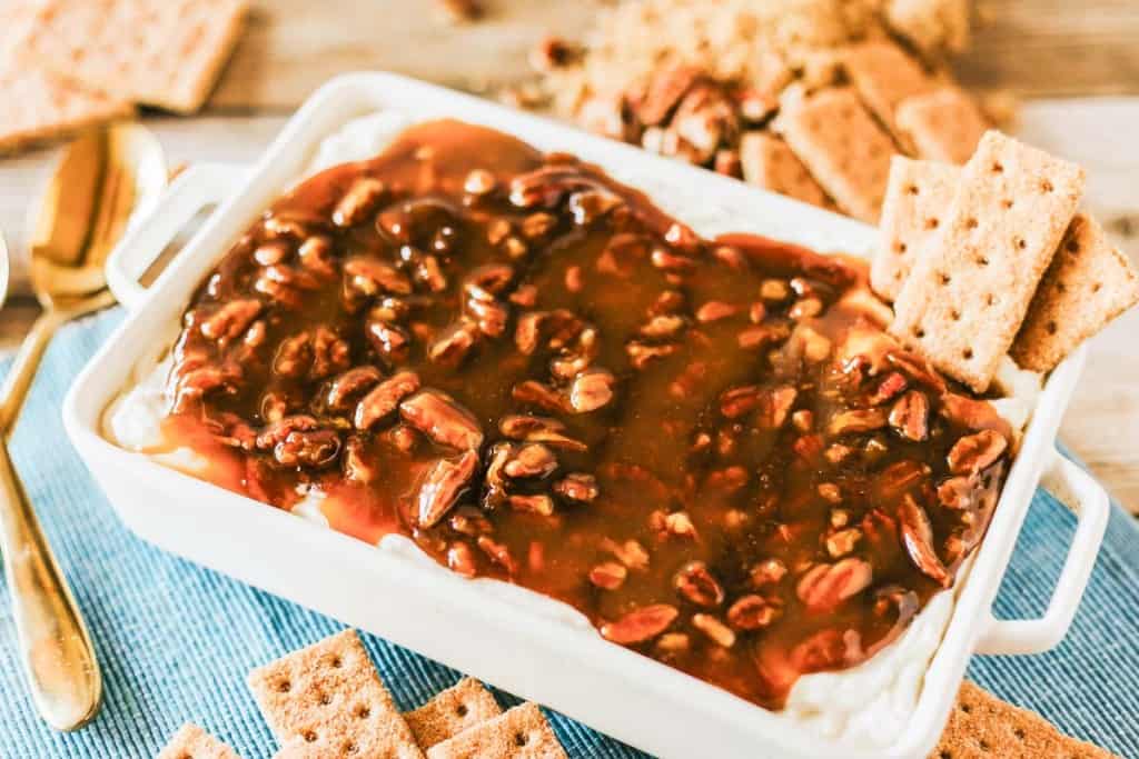 Rectangular dish filled with creamy pecan pie dip, topped with caramelized pecan sauce and surrounded by graham crackers on a blue cloth.