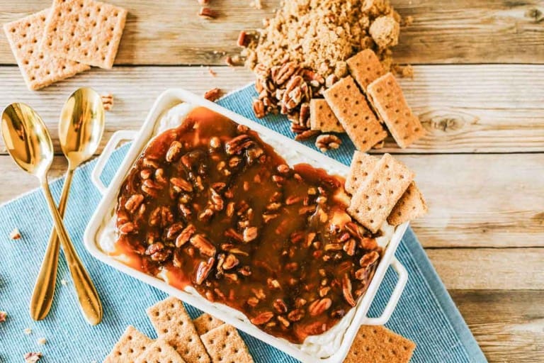 A rectangular dish filled with luscious pecan pie dip and topped with cream cheese dessert and praline sauce, surrounded by graham crackers, brown sugar, and two gold spoons on a blue cloth.