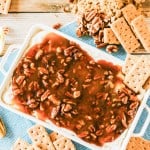 A rectangular dish of pecan pie dip with graham crackers on the side, surrounded by brown sugar, pecans, and more graham crackers on a blue cloth.