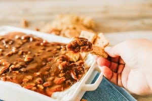 A hand dips a cracker into a baking dish filled with creamy, brown, chunky pecan pie dip on a wooden table.
