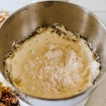 A metal mixing bowl contains creamed butter and sugar with dry ingredients on top, next to bowls of chopped pecans and brown sugar—perfect for starting a delicious pecan pie dip on a light wooden surface.
