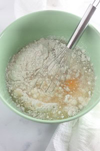 A green bowl containing flour, liquid ingredients, and a whisk, ready for mixing on a white surface with a white cloth nearby.