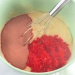 A green mixing bowl with cake batter, dry red powder, and finely chopped red gelatin, with a whisk resting inside.