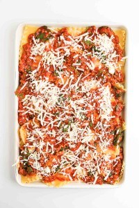 A baking dish filled with uncooked lasagna layers, topped with tomato meat sauce, spinach, and shredded cheese.