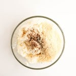A glass bowl containing ricotta cheese, grated parmesan, ground black pepper, and a drizzle of olive oil on a white surface.