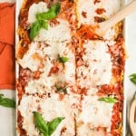 A rectangular baking dish filled with sliced lasagna topped with ricotta cheese, tomato sauce, and basil leaves, with a wooden spatula in one corner.